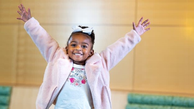 Children's Colorado patient - little girl wearing pink and a huge smile, holding her hands up in excitement and celebration