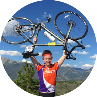 Smiling teen, Daniel, who overcame cancer, holding up his bike over his head triumphantly. He is wearing a red white and blue jersey, and the forested mountains and blue sky of the tour route area are seen in the background behind him